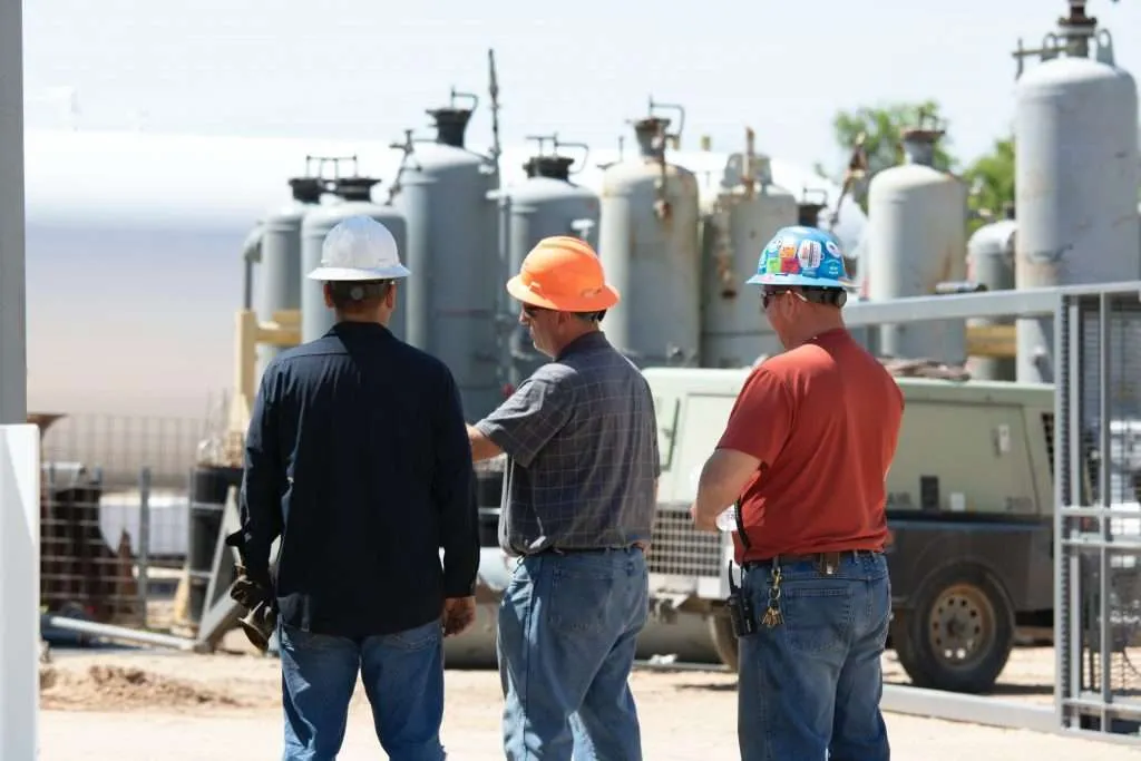 Three field workers in hard hats standing near industrial gas cylinders and a compressor unit