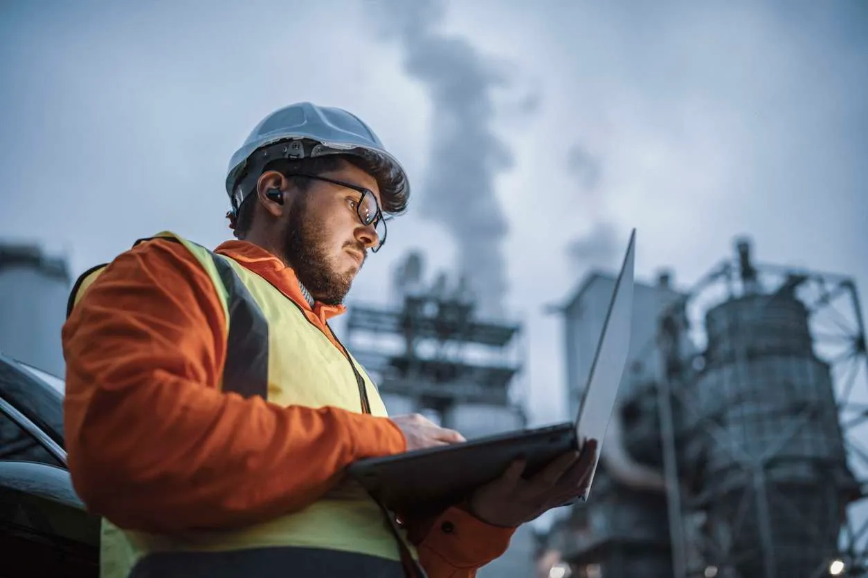 Male field service manager using a laptop while working in a factory - setting up prescriptive maintenance strategies for their assets.