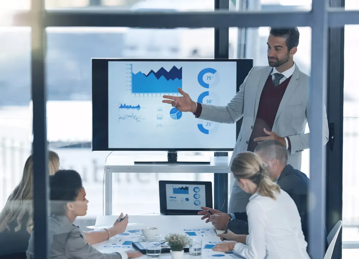 Presenter gesturing at charts and progress graphs on a screen during a meeting