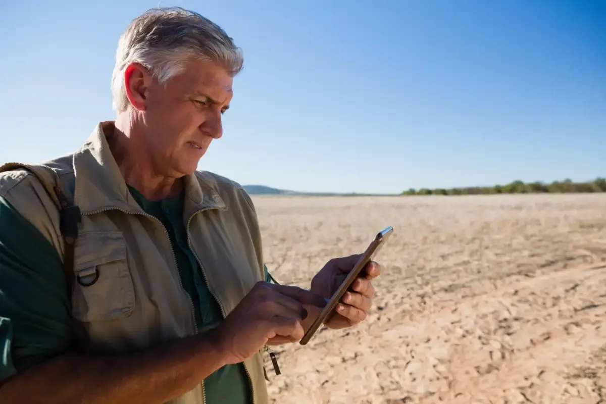 Man in a khaki vest using a handheld device while standing in a dry, cracked field