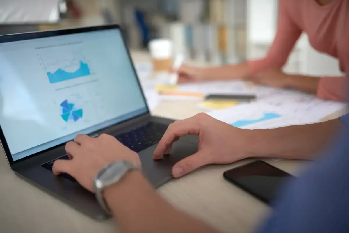 Laptop screen showing a line chart and a segmented data chart, with a colleague nearby and printed charts on the desk