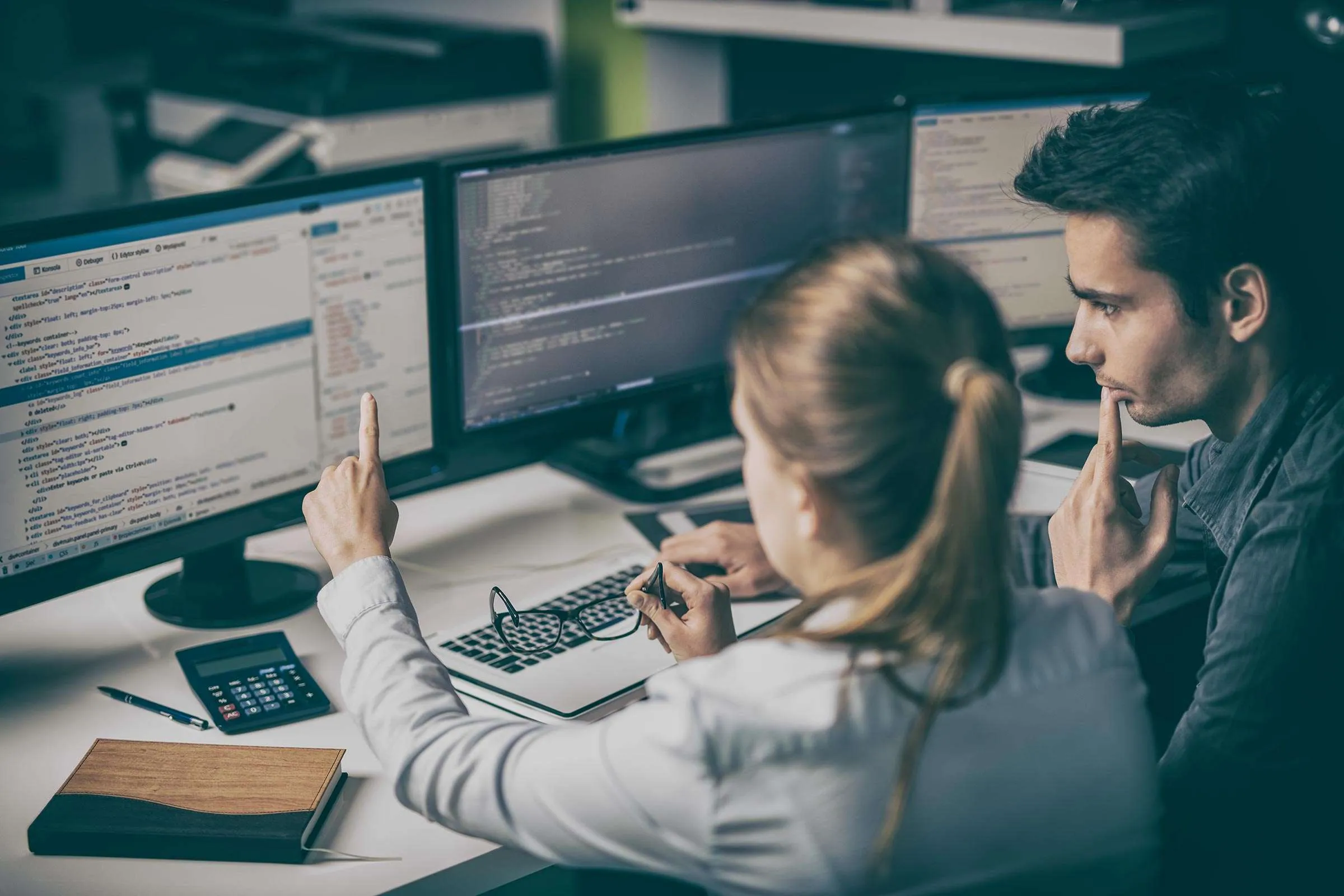Two developers reviewing code across three monitors, one pointing at the screen