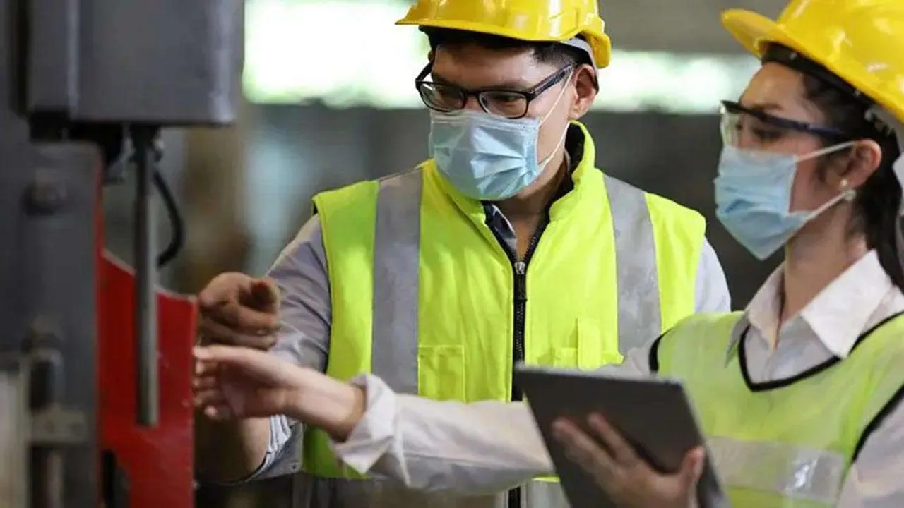 Two workers in hard hats, hi-vis and face masks inspecting machinery with a tablet