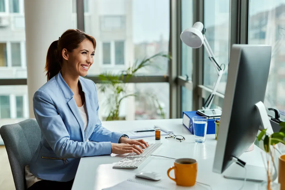 Smiling office worker typing at a desktop computer with a binder and coffee on her desk