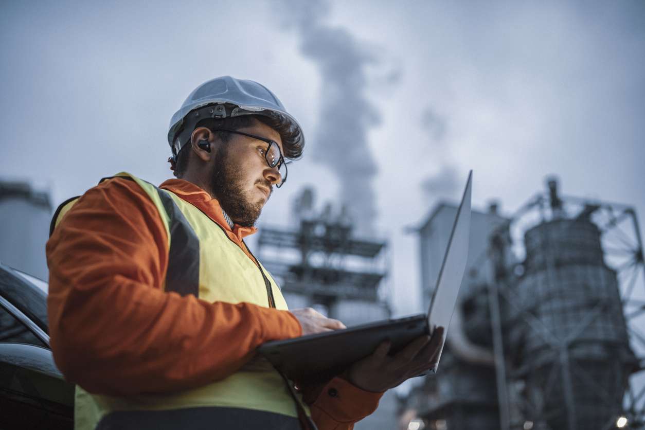 Male field service manager using a laptop while working in a factory - setting up prescriptive maintenance strategies for their assets.