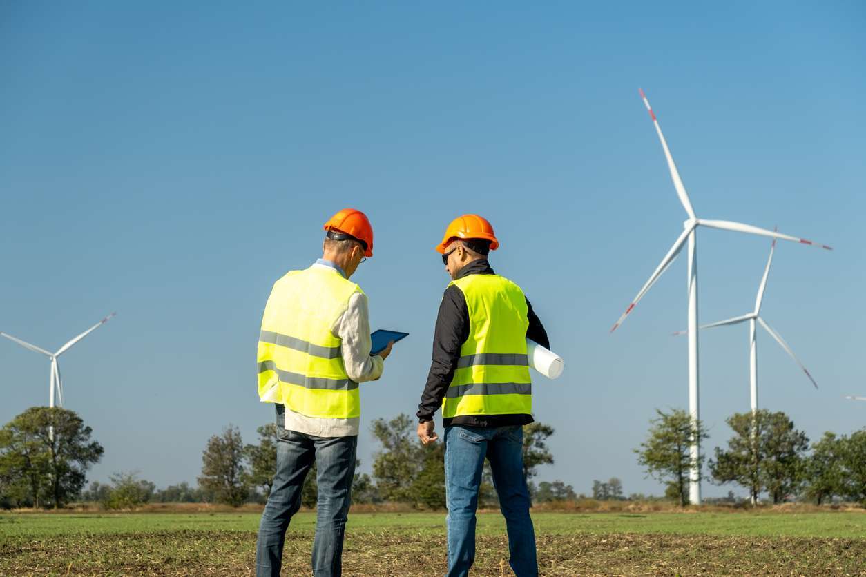 Environmental inspections - Male inspectors using a tablet device while checking windmills in a countryside.